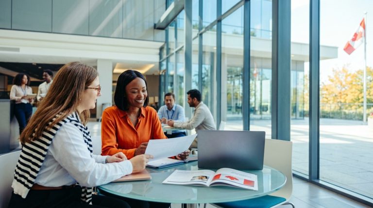 Two women review work permit documents in a Canadian office lobby as staff meet nearby